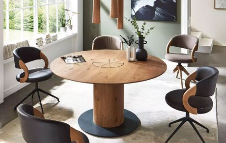 Ein Esstisch aus Holz mit einem runden Tischblatt und einem zylindrischen Sockel steht in einem Raum mit Stühlen. Auf dem Tisch stehen Vasen und Zeitschriften. Ein Fenster ist im Hintergrund.