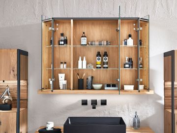 Offener Holz-Spiegelschrank mit Glasböden über einem Waschbecken. Darin Kosmetikartikel, Zahnbürsten, Seife und Flakons. Seitlich Holzschränke, graue Wand und schwarzer Waschtisch.