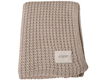Beige, grob gestrickte Decke, gefaltet. Ein helles Etikett mit dunkler Schrift "JOOP!" befindet sich auf der Decke. Die Decke hat ein offenes, löchriges Strickmuster.
