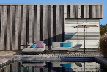 Helle Liegen mit Kissen auf Holzdeck am Pool, daneben eine Holzwand und ein Sonnenschirm. Reflexionen im Wasser.