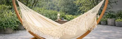 Eine weiße Hängematte mit Fransen hängt an einem hellbraunen Holzgestell. Im Hintergrund sind grüne Büsche und Bäume zu sehen, die einen ruhigen Garten andeuten.