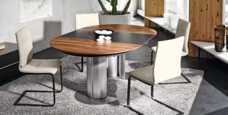 Ovaler Tisch mit Holz- und Metallelementen, umgeben von vier Stühlen mit hellem Lederbezug und schwarzem Gestell. Dekoschale in der Mitte. Teppich. Pflanze im Hintergrund.