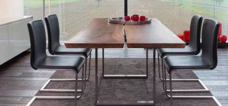 Ein Esstisch aus zwei Holzplatten mit vier Stühlen aus dunklem Leder und Metallbeinen steht auf einem dunklen Teppich. Auf dem Tisch eine silberne Schale mit roten Deko-Objekten.