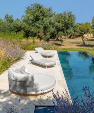 Ein Poolbereich mit rundem Sofa, zwei Liegen und blühenden Pflanzen am Beckenrand. Im Hintergrund Bäume und blauer Himmel.