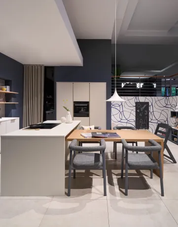 Cuisine ouverte moderne avec coin repas, tons bleu foncé et beige clair. Îlot avec plaque de cuisson et poêle face à une table en bois, chaises grises et chaise haute enfant. Suspension blanche. Mur bleu foncé avec lignes abstraites et tableau noir.