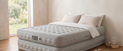 Graues Intex Luftbett mit strukturierter Oberfläche und eingebautem Bedienfeld in hellem Zimmer auf Holzboden. Bezogen mit weißer Bettwäsche und zwei hellbeigen Kissen. Großes Fenster zeigt verschwommene grüne Landschaft.