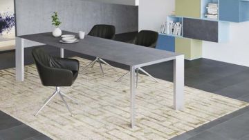 Ein grauer Esstisch mit weißen Beinen steht auf einem gemusterten Teppich. Drei schwarze Stühle stehen um den Tisch. Auf dem Tisch stehen Geschirr, eine Tasse und eine kleine Pflanze in einer weißen Vase.