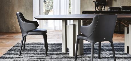 Esstisch mit dunkler Platte und hellen, würfelförmigen Beinen. Zwei schwarze Stühle mit Armlehnen auf einem gemusterten Teppich. Im Hintergrund ein Fenster und eine Kommode mit Dekoration.