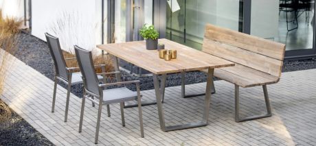 Ein Esstisch aus Holz und Metall steht mit einer Holzbank und zwei Stühlen auf einer Terrasse. Dekorationen wie Pflanzen und Kerzen sind auf dem Tisch.