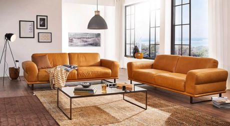 Wohnzimmer mit zwei braunen Ledersofas, Couchtisch, Teppich und großem Fenster mit Ausblick. An der Wand hängen Bilder. Eine Stehlampe und eine Hängelampe beleuchten den Raum.