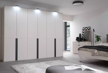 Ein helles Schlafzimmer mit großem, weißem Kleiderschrank, Bett und Kommode. Ein grauer Teppich und Hocker ergänzen den Raum. Dekorative Elemente wie Pflanzen und Bilder runden das Ambiente ab.