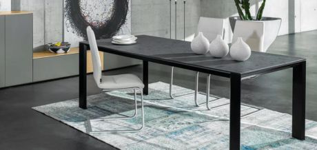 Ein Esstisch mit Stühlen steht auf einem Teppich, darauf Teller und Vasen. Im Hintergrund eine moderne Dekoration und ein Schrank.