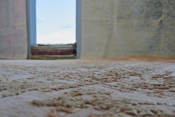 Nahaufnahme eines beigen Teppichs mit erhabenem Muster. Im Hintergrund unscharf ein Fenster mit blauem Himmel und weitere Teppiche.