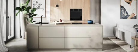 Cuisine ouverte moderne avec îlot central gris clair, évier noir et plaque à induction avec casserole bleue. Derrière, rangements muraux en bois clair et four encastré noir. À gauche, plante verte et poster. À droite, canapé et tapis. Ombres longues.