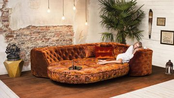 Innenaufnahme eines Wohnzimmers mit orangefarbenem Sofa, auf dem eine Frau liegt. Pflanze, Dekorationen und Beleuchtung ergänzen den Raum.