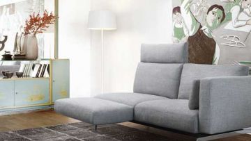 Helles Wohnzimmer mit grauer Couch, Stehlampe und blau-goldenem Schrank. An der Wand hängt ein abstraktes Gemälde. Ein dunkler Teppich liegt auf dem Holzboden.