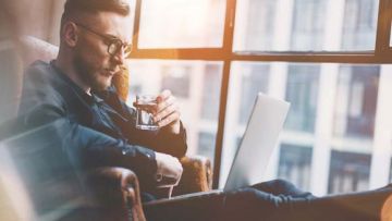 Ein Mann mit Brille sitzt im Sessel, hält ein Glas mit Flüssigkeit und arbeitet am Laptop. Im Hintergrund ist ein Fenster mit Blick auf ein Gebäude.