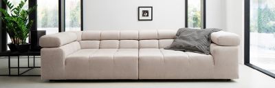 Ein beiges modulares Sofa steht in einem hellen Raum. Ein schwarz-weiß karierter Überwurf liegt auf einem Ende. Pflanzen und ein Tisch stehen links, große Fenster mit schwarzem Rahmen im Hintergrund.