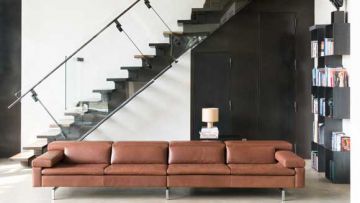 Ein Wohnraum mit einer braunen Ledercouch, einer Treppe mit Glasgeländer, einer Lampe auf einem Tisch und einem Bücherregal. Die Wand ist weiß und dunkel.