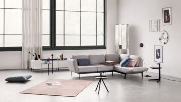 Ein Wohnzimmer mit großem Fenster, grauer Couch, Kissen, Teppich, Lampen, Tischchen und Deko. Die Wände sind weiß, der Boden grau. Das Fenster hat schwarze Rahmen.