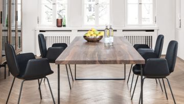 Ein Esstisch aus Holz mit sechs schwarzen Stühlen in einem hellen Raum mit Parkettboden. Auf dem Tisch stehen eine Schale mit Obst und Wasserflaschen.
