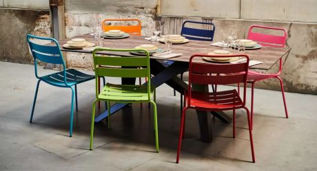 Esstisch mit sechs bunten Stühlen. Auf dem Tisch sind Teller, Gläser und Besteck für sechs Personen angeordnet. Der Tisch steht in einem Raum mit Betonwänden.