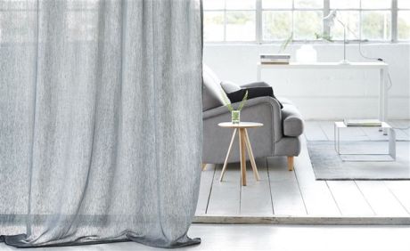 Ein Wohnraum mit einem grauen Vorhang, einem grauen Sofa, einem Holztisch und weißen Möbeln. Helles Licht dringt durch die Fenster.