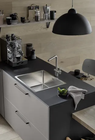Kitchenette avec plan de travail foncé et placards gris clair. À gauche, une machine à expresso ; au centre, un évier avec un robinet ; à droite, un bol contenant des poivrons verts et un torchon. Suspension noire.