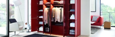 Einrichtung mit offenem, rotem Kleiderschrank, der Kleidung und Handtücher zeigt. Ein Sessel und eine Stehlampe stehen in der Nähe des Fensters. Eine rote Couch steht im Hintergrund.