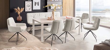 Ein Esszimmer mit einem weißen Tisch, umgeben von grauen Stühlen mit schwarzen Beinen. Eine Bank mit Kissen steht an einer Seite des Tisches. Im Hintergrund ein Fenster mit Meerblick.