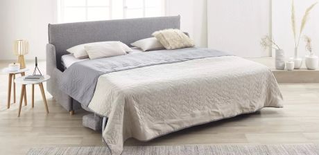 Ein graues Sofa mit Bettfunktion ist mit Kissen und Decken in neutralen Farben dekoriert. Daneben stehen Beistelltische mit Deko und Vasen.