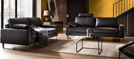 Wohnzimmer mit dunklem Ledersofa und Sessel auf einem Teppich. Ein Couchtisch steht davor. Im Hintergrund sind eine gelbe Wand und eine Treppe zu sehen.