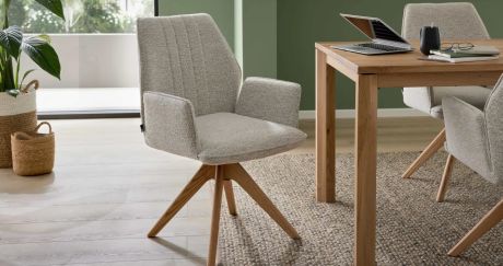 Einrichtungsszene mit beigefarbenem Polsterstuhl auf Holzbeinen neben Holztisch. Auf Tisch Laptop, Tasse, Brille und Notizbücher. Im Hintergrund Zimmerpflanze und Fenster.