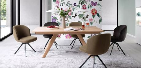 Ein Esszimmer mit Holztisch und Stühlen. Auf dem Tisch stehen Blumen, eine Schale mit Beeren, ein Buch, Wein und Gläser. Im Hintergrund eine Wand mit floralem Muster.