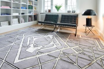 Wohnzimmer mit hellem Holzboden, Teppich mit geometrischen Mustern in Grau und Weiß, zwei schwarzen Lederstühlen und einem Bücherregal im Hintergrund.
