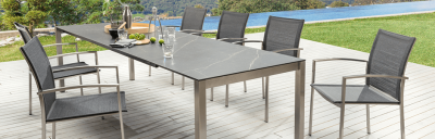 Esstisch mit grauer Platte und sechs Stühlen auf einer Terrasse. Auf dem Tisch stehen Getränke und Dekoartikel. Im Hintergrund sind ein Pool und grüne Vegetation zu sehen.