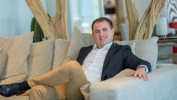 Ein Mann lächelt, während er lässig auf einem hellen Sofa liegt, mit beigen Hosen, schwarzem Sakko und Schuhen, die einen entspannten, aber stilvollen Eindruck vermitteln.