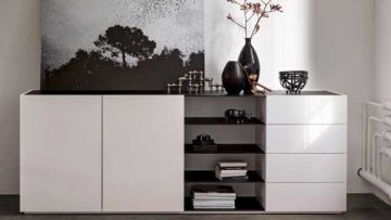 Weißes Sideboard mit schwarzen Akzenten, darauf Deko wie Vasen, Skulpturen, Bücher und ein Schwarzweiß-Bild mit Baum-Motiv. Seitlich ein weißer Heizkörper.