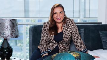 Eine lächelnde Frau sitzt auf einem Bett vor einem Fenster. Sie trägt eine gemusterte Jacke und eine Halskette mit einer Perle. Neben ihr steht eine Lampe mit einem dunklen Lampenschirm.