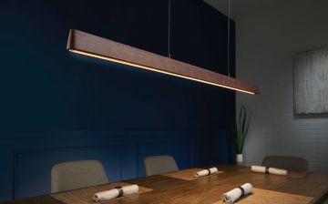 Esstisch mit Gedecken unter einer hängenden, länglichen Holzlampe vor einer blau-grauen Wand. Pflanzen und ein Bild sind im Hintergrund erkennbar.