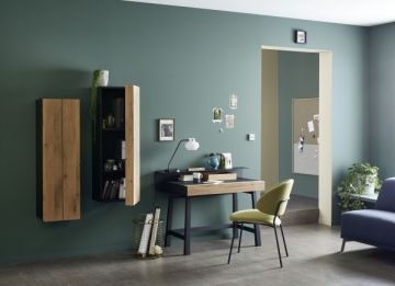 Ein Arbeitszimmer mit grün gestrichenen Wänden. Ein Schreibtisch mit einem gelben Stuhl davor. An der Wand hängen Holzregale und Fotos. Im Hintergrund ist ein Durchgang zu einem anderen Raum sichtbar.