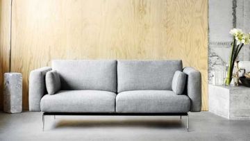 Graues Sofa mit Kissen vor hellbrauner Holzvertäfelung. Rechts davon ein Betonsockel mit Blumen und Dekoration. Links ein grauer, quaderförmiger Hocker.