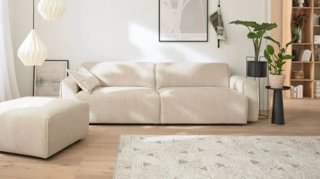 Ein helles Wohnzimmer mit beiger Couch, passendem Ottoman, gemustertem Teppich und Pflanzen. Helle Holzböden und weiße Wände tragen zur freundlichen Atmosphäre bei. Moderne Lampen hängen von der Decke.