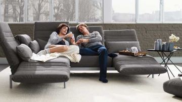 Ein Paar entspannt auf einem grauen Sofa mit verstellbaren Rückenlehnen vor einem Fenster. Ein Tisch mit Getränken und Kerzen steht daneben.