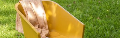 Gelber Sessel im Freien auf Rasen, mit beiger Decke drapiert. Das Sonnenlicht scheint auf die Sitzfläche.