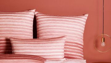 Nahaufnahme von Bettwäsche mit horizontalen Streifen in Rosa- und Weißtönen. Eine goldene Lampe hängt rechts im Bild vor einer rosa Wand.