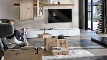 Wohnzimmer mit modernem Mobiliar: helles Holzregal, weisses Lowboard unter einem Fernseher. Eine Frau sitzt im Sessel. Ein ungewöhnlicher Couchtisch aus Holzblöcken steht auf einem hellen Teppich.