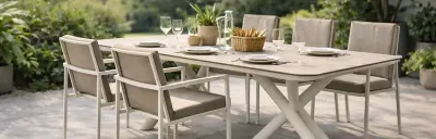 Ein Esstisch mit sechs Stühlen im Freien. Der Tisch ist gedeckt mit Tellern, Gläsern und einer Brotkorb. Pflanzen und Bäume sind im Hintergrund zu sehen.