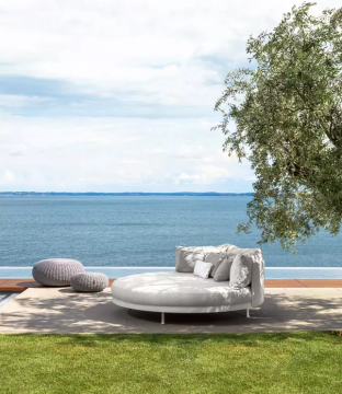 Rundes, weißes Daybed mit Kissen und zwei Poufs auf einer Terrasse mit Meerblick unter einem Baum.