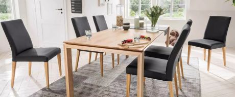 Ein Esstisch aus hellem Holz steht im Raum, umgeben von sechs Stühlen mit dunklen Bezügen und hellen Holzbeinen. Auf dem Tisch stehen Dekorationen wie Blumen und Spielzeug.